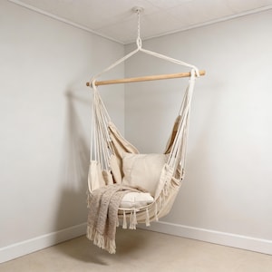 Può includere: Una sedia sospesa beige con una barra di legno e supporti in corda bianca. La sedia ha un cuscino beige e una coperta abbinata con frange. La sedia è sospesa al soffitto in un angolo della stanza.