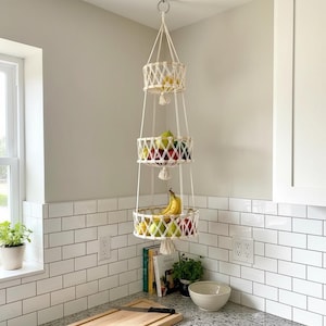 Puede incluir: Una cesta de frutas colgante de macramé de tres niveles llena de varias frutas. Las cestas blancas tejidas están suspendidas de un anillo de metal. El entorno de la cocina presenta azulejos blancos de metro y una tabla de cortar de madera.
