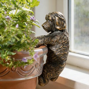 May include: A bronze-colored dog sculpture clinging to the edge of a terracotta flower pot. The dog has detailed fur and is positioned as if looking at the plant. The pot contains green foliage and small purple flowers.