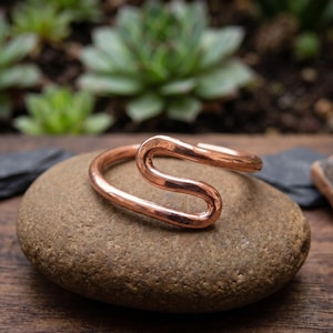 Pode incluir: Um anel de cobre polido com um design fluido e contínuo em forma de S. O metal tem um tom castanho-avermelhado metálico quente com uma superfície lisa e reflexiva. O anel é formado por uma única peça de fio arredondado que se curva numa forma aberta.