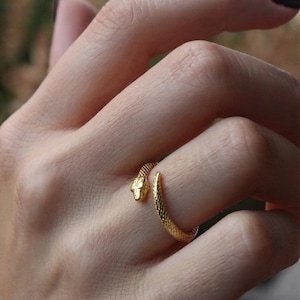 Può includere: Un anello aperto color oro a forma di serpente avvolto. La fascia presenta una texture a scaglie su tutta la superficie, con una testa definita a un'estremità e una coda affusolata all'altra, che si avvolge attorno al dito in una spirale.