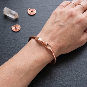 Könnte beinhalten: Ein polierter Kupfer-Armreif mit einer gedrehten, gehämmerten Textur. Der Armreif weist ein durchgehendes, wellenförmiges Design auf, das sich um das Handgelenk legt. Die Metalloberfläche reflektiert das Licht und betont die unregelmäßige, handgefertigte Optik.