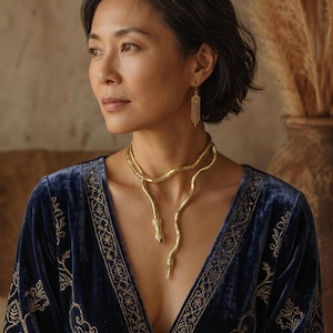 Puede incluir: Un collar dorado con forma de serpiente que se envuelve alrededor del cuello con dos extremos colgantes, combinado con un pendiente dorado con una pieza geométrica pequeña y un colgante de flecos de varias cadenas. Usado con un top de terciopelo azul.