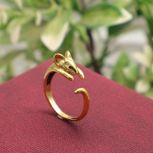 May include: A gold-colored ring featuring a mouse design. The ring wraps around the finger with the mouse's head and paws on top. The ring is set against a red background.