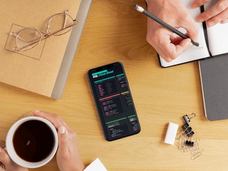 K&ouml;nnte beinhalten: Draufsicht auf einen Holztisch mit einem Smartphone, das ein SQL-Cheat-Sheet anzeigt. Eine Brille, ein Notizbuch und eine Kaffeetasse befinden sich ebenfalls auf dem Tisch. Eine Person schreibt mit einem Bleistift in ein Notizbuch.