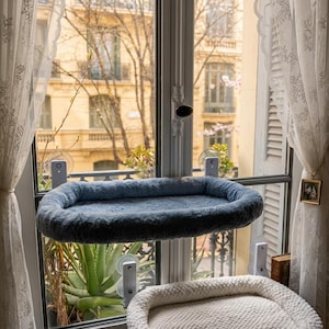 Puede incluir: Una cama para gatos azul de forma ovalada, suspendida de una ventana con ventosas. La cama tiene una textura suave y afelpada. Debajo se ve una segunda cama para gatos blanca. La ventana da a una calle de la ciudad.