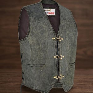 Könnte beinhalten: Eine Weste aus grauem Leder im Used-Look mit V-Ausschnitt und drei messingfarbenen Verschlüssen. Die Weste hat zwei Vordertaschen und dekorative Nähte an den Kanten. Das Etikett lautet "MANRACING".
