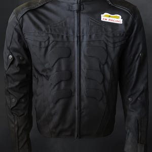 May include: Black motorcycle jacket with a stand-up collar and zip closure. The jacket has padded sections on the chest and shoulders, and a small patch that reads "FM RACING". It features multiple pockets and button details.