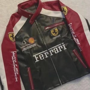 May include: Black, red, and white Ferrari racing jacket. The jacket features the Ferrari logo on the chest and sleeves, with the Ferrari name prominently displayed across the front. It has a zip-up front, red zippers, and a stand-up collar.