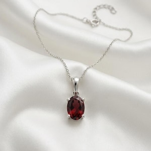 Op de afbeelding: Een zilveren ketting met een hanger met een grote, ovaal geslepen, dieprode edelsteen. De hanger is gezet in een zilveren rand en hangt aan een fijne zilveren ketting. De achtergrond is een witte, zijdeachtige stof.