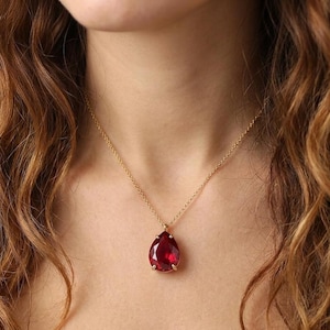 May include: A gold necklace featuring a large, teardrop-shaped, red gemstone pendant. The pendant is set in a gold bezel and hangs from a delicate gold chain. The necklace is displayed against a skin tone background.