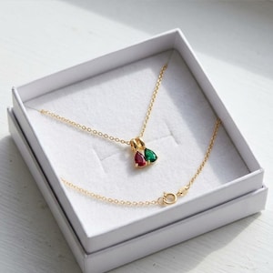 Può includere: Una collana dorata con un ciondolo con due pietre preziose a forma di goccia, una rossa e una verde, presentata in un cofanetto bianco. La catena della collana è delicata e la chiusura è visibile.