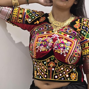 Può includere: Top ricamato vivace con base rossa e nera, ornato da colorati motivi geometrici, specchi e motivi floreali. Le maniche hanno un bordo metallico. È visibile anche una collana d'oro.