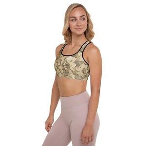 May include: A beige sports bra with a black trim and a camouflage pattern. The bra has a scoop neckline and a racerback design. The model is wearing pink leggings.