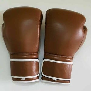 Puede incluir: Un par de guantes de boxeo de cuero marrón. Los guantes tienen correas blancas para la muñeca y costuras blancas. Están diseñados para la protección y la absorción de impactos.
