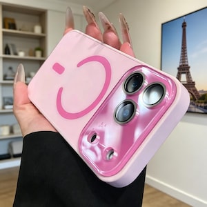 May include: A light pink phone case with a bright pink smiley face design. The case has a clear, glossy camera lens protector. The phone case is held in a person's hand.