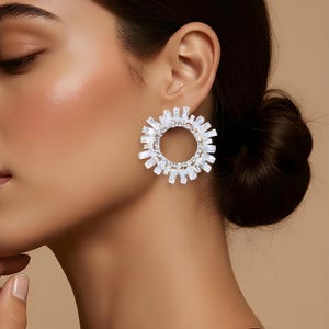 Può includere: Un paio di orecchini circolari scintillanti. Ogni orecchino presenta un anello centrale circondato da un alone di cristalli rettangolari trasparenti. Gli orecchini sono posti su uno sfondo neutro, evidenziando la loro lucentezza e il design.
