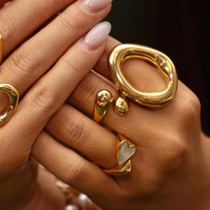 Puede incluir: Primer plano de anillos dorados con diferentes diseños. Uno tiene forma ovalada grande, otros tienen forma de lágrima o corazón. Los anillos se muestran en una mano con uñas cuidadas, mostrando las joyas.