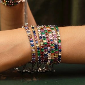 May include: A collection of colorful gemstone bracelets. The bracelets feature a variety of rectangular-cut stones in shades of red, blue, green, yellow, and purple. The bracelets are silver-toned and appear to be adjustable.