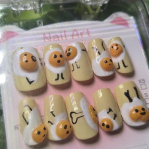 Può includere: Un set di dieci unghie finte con un design a uovo a cartoni animati. Ogni unghia è dipinta di giallo con un bianco d'uovo bianco e un tuorlo arancione sorridente. Linee e dettagli neri completano il design dell'uovo. Le unghie sono in una confezione di plastica trasparente.