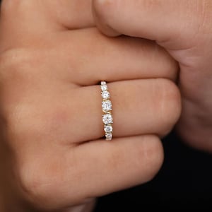 May include: A gold ring with a series of graduated, round-cut diamonds. The ring showcases multiple clear, sparkling stones, creating a delicate and elegant design. The ring is displayed on a person's finger against a dark background.