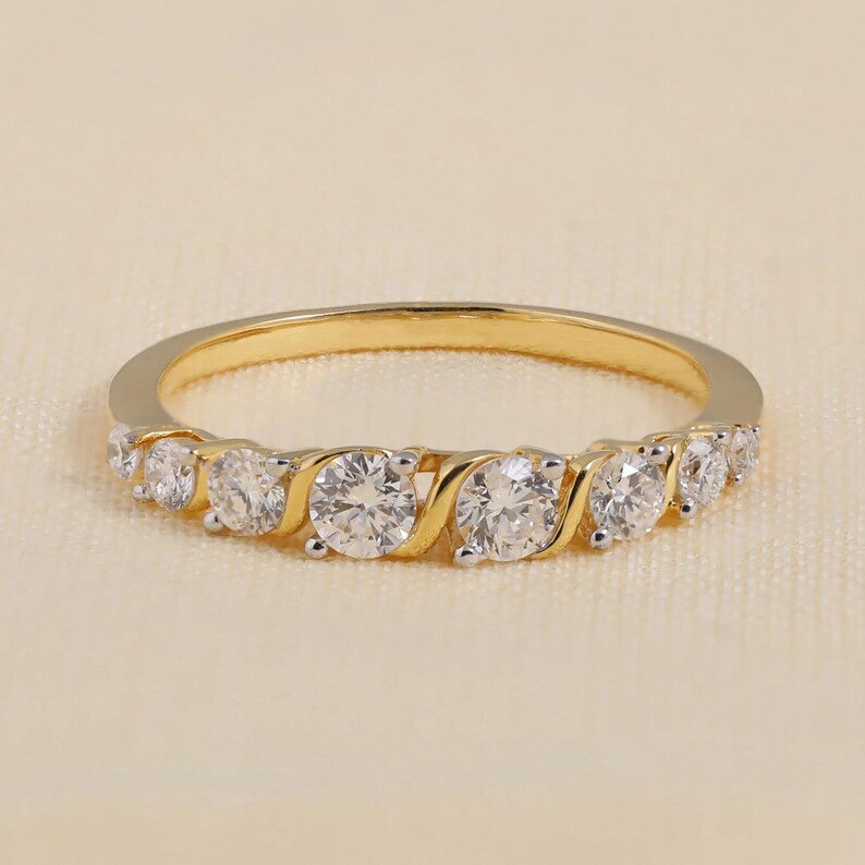 May include: A gold ring featuring seven clear, round gemstones. The stones are set in a swirling design, interspersed with gold sections. The ring is presented on a cream-coloured surface.