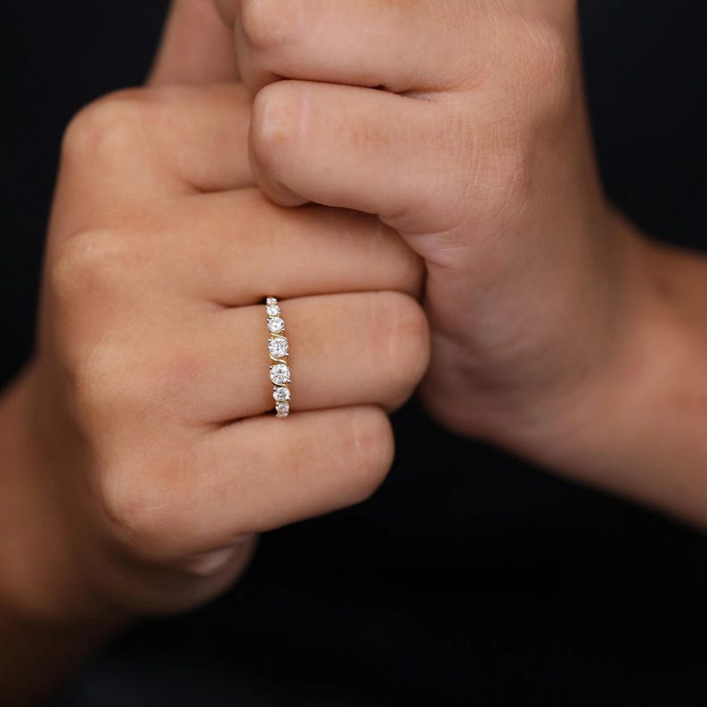 May include: A gold ring with a series of graduated, round-cut diamonds. The ring showcases multiple clear, sparkling stones, creating a delicate and elegant design. The ring is displayed on a person's finger against a dark background.
