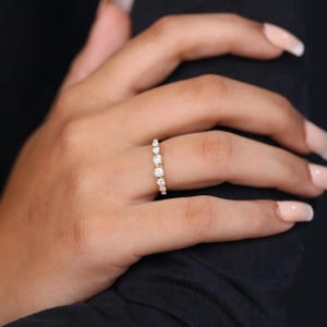 May include: A gold ring featuring a series of clear, round gemstones. The ring is worn on a finger, set against a dark backdrop. The gemstones are of varying sizes, with the larger ones positioned centrally.