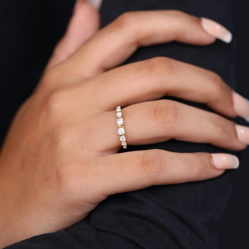 May include: A gold ring featuring a series of clear, round gemstones. The ring is worn on a finger, set against a dark backdrop. The gemstones are of varying sizes, with the larger ones positioned centrally.
