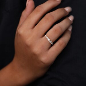 May include: A delicate gold ring with a series of small, round, clear gemstones. The ring has gemstones graduating in size, creating a subtle sparkle. The ring is shown on a hand against a dark background.