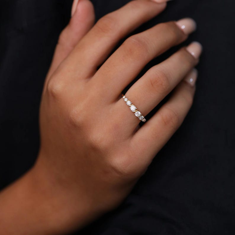 May include: A delicate gold ring with a series of small, round, clear gemstones. The ring has gemstones graduating in size, creating a subtle sparkle. The ring is shown on a hand against a dark background.