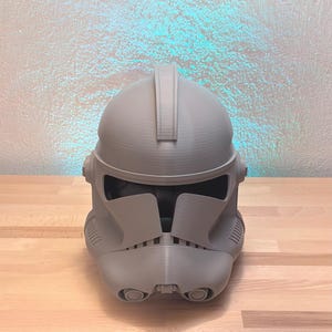 May include: A gray 3D printed clone trooper helmet featuring a smooth dome, a central ridge, a wide horizontal visor opening, and a detailed mouth section with two circular vents. The helmet has a matte finish and visible layer lines from the printing process.
