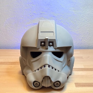 Könnte beinhalten: Ein hellgrauer, 3D-gedruckter AT-AT-Pilotenhelm. Der Helm hat ein unverwechselbares Design mit Visier, Atemschutzgerät und verschiedenen mechanischen Details. Der Helm ist zum Tragen bestimmt und ist eine Replik aus einem Science-Fiction-Film.