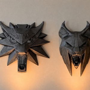 Può includere: Due sculture da parete geometriche grigio scuro che raffigurano teste di lupo stilizzate con tratti angolari e bocche aperte che mostrano i denti. La scultura a sinistra ha una criniera larga e appuntita, quella a destra orecchie alte e appuntite.