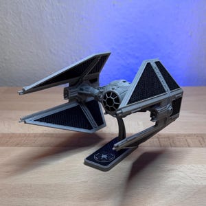 Könnte beinhalten: Ein graues TIE-Interceptor-Modell eines Sternenjägers mit vier abgewinkelten Solarflügeln mit schwarzen, texturierten Paneelen, einem zentralen kugelförmigen Cockpit mit rundem Sichtfenster und einem Ständer mit imperialem Emblem.