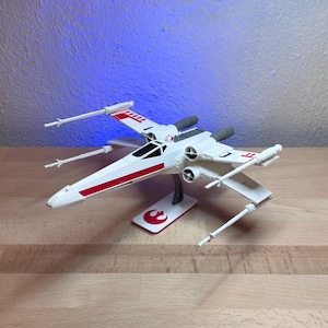 Pode incluir: Um modelo de nave espacial X-wing branca com marcações vermelhas nas asas e uma cabine escura. A nave está montada num suporte branco com o símbolo vermelho da Aliança Rebelde, quatro canhões laser e dois motores na parte traseira.