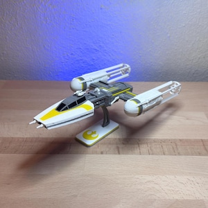 Puede incluir: Un modelo de caza estelar Y-wing blanco y gris con detalles amarillos en el morro y los motores. La nave espacial cuenta con una pequeña figura de droide detrás de la cabina y está montada sobre una base con un símbolo rebelde amarillo.