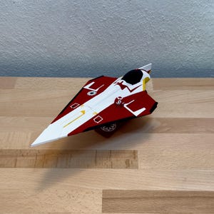 May include: A red, white, and black model starfighter with a sleek, angular design. The model features a detailed cockpit, wing markings, and a central emblem. The starfighter is resting on a wooden surface.