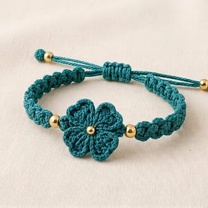 May include: Teal crochet bracelet with a flower motif at the centre. It features gold-coloured beads and an adjustable sliding knot fastening. The flower and bracelet are made of teal yarn.
