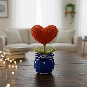 May include: A handmade crochet flower with a heart-shaped orange bloom, green leaves, and a blue pot. The pot is decorated with white hearts and a wavy pattern. The flower sits on a dark wooden table.