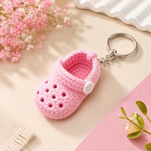 May include: A light pink crochet keychain shaped like a clog shoe. The miniature footwear features a textured knit pattern with perforated holes on the toe area, a pivoting heel strap, and a small white circular button on the side attached to a ring.