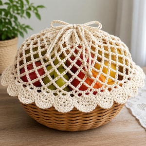 May include: A round, light brown woven wicker basket filled with red apples, green grapes, and oranges. The basket is covered by a cream-colored, crocheted net fabric with a scalloped edge and a drawstring top secured with two small wooden beads.