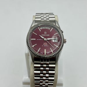 Peut inclure: Une montre-bracelet automatique Ricoh argentée avec un cadran bordeaux. La montre présente un affichage du jour et de la date, une lunette cannelée et un bracelet en acier inoxydable. Le cadran affiche le mot "Wednesday" et "21 Jewels".