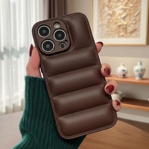 May include: A brown, quilted phone case with a camera cutout. The case has a padded, textured appearance with horizontal stitching. The case is designed to fit a smartphone and offers protection. The case is held by a person wearing a green sweater.