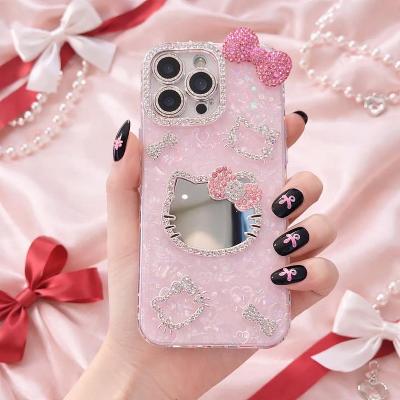 May include: Pink phone case with a Hello Kitty design. The case features a mirror, a bow, and sparkling embellishments. The case has a glittery texture and is decorated with rhinestones. The phone case is a cute accessory.