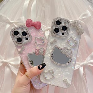 May include: Two decorative phone cases. One is pink, the other is white. Both cases feature a Hello Kitty design with bow accents and a sparkling, jeweled border around the camera. The cases are embellished with rhinestones.