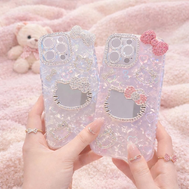 May include: Two clear phone cases with a sparkly, iridescent finish. Each case features a Hello Kitty design with a bow and other decorative elements made of rhinestones. The cases are held up by a person's hands, and the background is a soft pink.