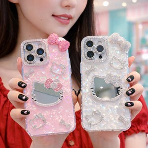 May include: Two decorative phone cases, one pink and one white, featuring a Hello Kitty design. Both cases are embellished with rhinestones and bows. The pink case has a mirrored Hello Kitty face and a pink bow on top.