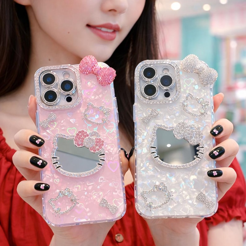 May include: Two decorative phone cases, one pink and one white, featuring a Hello Kitty design. Both cases are embellished with rhinestones and bows. The pink case has a mirrored Hello Kitty face and a pink bow on top.