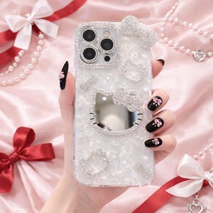 May include: A clear phone case with a pearlescent finish, adorned with sparkling rhinestones. The case features a bow, a Hello Kitty design, and other decorative elements. The phone case is held by a hand with black nail polish and pink flower accents.